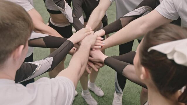 High angle cropped shot of team of cheerleaders stacking hands, then raising arms up and smiling while huddling on football field before match