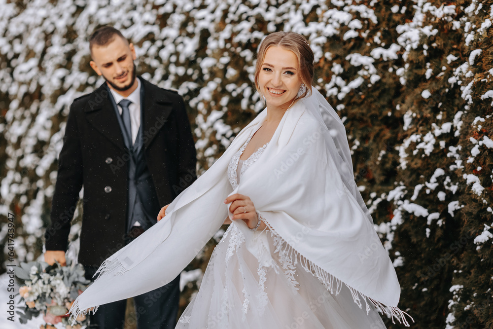 Wall mural Portrait of happy newlyweds on the background of snow-covered trees. The groom hugs the bride in the winter park. Smiling bride in wedding dress and white poncho. The groom is dressed in a black coat.