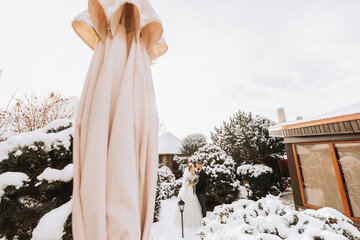 happy newlyweds between snowy trees. The groom hugs the bride in the winter park. Smiling bride with a bouquet of flowers in a wedding dress and white poncho. The groom is dressed in a black coat.