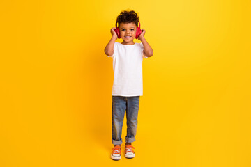 Full length photo of positive cheerful small boy wear white t-shirt listening music headphones isolated yellow color background