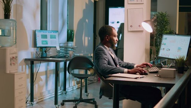 Dolly in shot of adept computer operator sitting at desk, looking over statistics charts, imputing data during nightshift. Startup staff personnel solving project task at work overnight