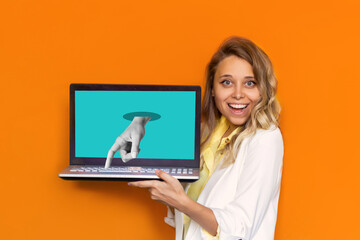 Young attractive smiling blonde woman in a white jacket showing laptop with turquoise screen with statue's hand pressing the keyboard key isolated on a bright orange color background. Modern design