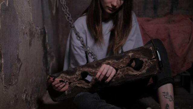 A young woman is sitting on the floor chained to antique wooden shackles with a chain in a dark room. Quest is a room with creepy tasks for adults. A room of fear or horror.