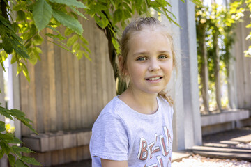 Portrait of a cute and pretty blonde little girl against a background of green broad-leaved plants....