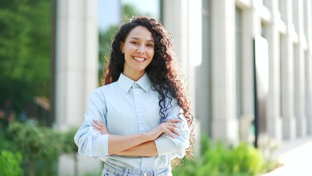 Portrait of a young smiling positive woman with crossed arms standing on the street near an office building. Head shot of a happy cheerful curly brunette female employee or manager looking at camera