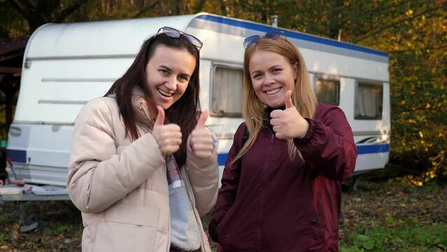 Portrait of two adult women In the woods against the background of a van in autumn, they look into the camera and give a thumbs up.