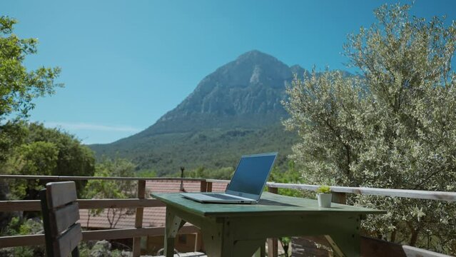 remote freelancer workplace with laptop on table outdoors on wooden terrace in nature. campsite and bungalow with terrace in amazing place with view on mount, hills and green woods at sunny day.