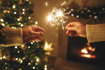 Happy New Year! Hands holding burning fireworks against modern fireplace and christmas tree with...