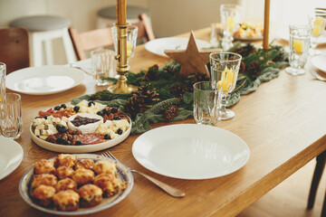 Christmas table setting. Stylish table runner with fir branches and pine cones, candles, cheese appetizers and starters, glasses and cutlery. Holiday dinner arrangement. Happy new year!