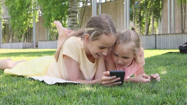 Two cute blonde little girls are relaxing in the park on the grass, watching videos or social networks, sending messages using a smartphone. Affordable wireless internet for teenagers.