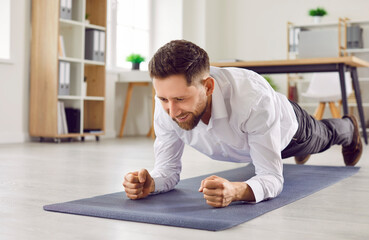 Portrait of a young cheerful business man doing plank exercise on yoga mat on the floor in office....