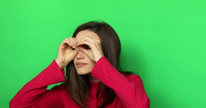 Amazed curious woman with brunette hair in sweater looking around through fingers imitating binoculars and point at something surprised. Indoor studio shot isolated on green background.