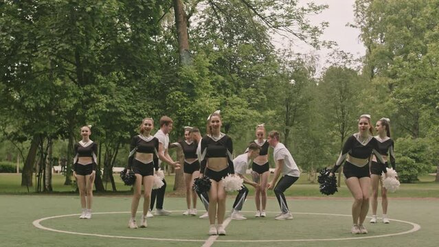 Wide shot of group of cheerleaders dancing with pompoms and performing partner stunts on outdoor soccer field