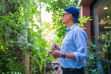 Asian man in blue shirt, hat and sunglasses, holding tablet to work in park with green plants.