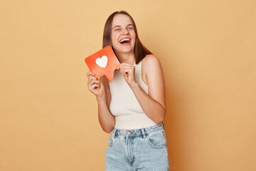 Blog communication. Blogger network. Web communication. Like counter. Laughing brown haired young woman wearing casual clothing holding red like icon standing isolated over beige background