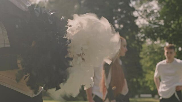 Cropped shot of cheer girl standing outdoors on field and shaking black and while pompoms at sunny summer day