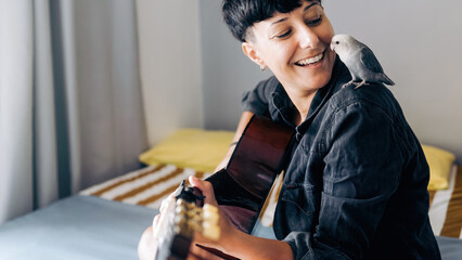 Happy woman and parrot bird enjoying together with guitar at home - Owner and pet relationship, love concept - obrazy, fototapety, plakaty