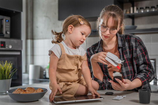 One young woman playing with toddler girl in the kitchen with finger puppets and holding tablet, babysitting at home concept