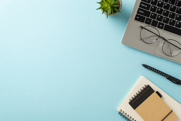 Business office atmosphere. Top view of laptop, notebooks, pen, potted plant, glasses on pastel blue setting. Empty area for text or advertorial content