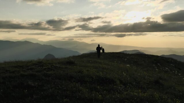 Silhouette of a man walking on top of a mountain with a rucksack at sunset. The wind is ruffling the grass. In the background silhouettes of mountain peaks. Epic video of mountain tourism