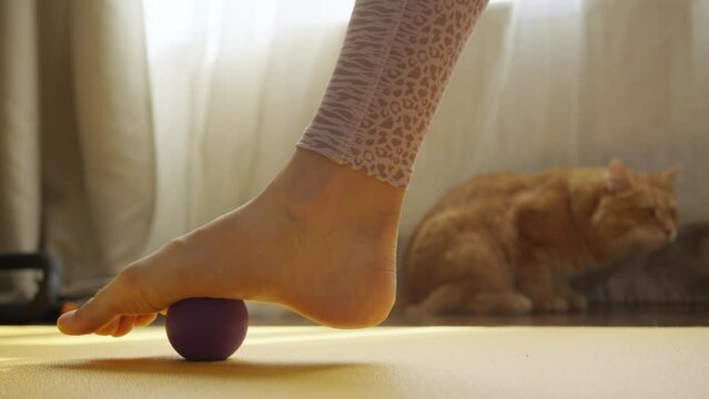 A girl kneads her foot with a massage ball after a sports injury at home on a sports mat close-up