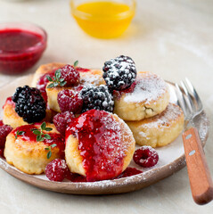 Syrniki serving in a ceramic plate, with raspberries and blackberries, sprinkled with honey and raspberry jam, sprinkled with powdered sugar, horizontal.
