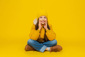 A young girl is sitting on the floor in full height, cross-legged, in autumn warm clothes. A child in a yellow warm jacket and a long beanie hat. Clothes for children. Yellow isolated background.