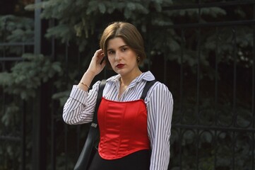 Fototapeta premium A young girl in a fashionable white shirt, red top and black skirt stands near a wall entwined with green leaves and straightens her hair.