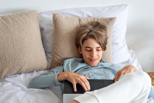 Hendsome teenager boy laying on bed with laptop, communicating online in social media, chatting, websurfing. Leisure pastime, youth technology addiction. Child with digital gadget