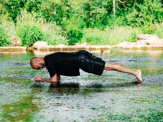 Man doing plank exercise in a water of a small stream. Warm sunny day. Unusual and exotic place to do workout. Healthy lifestyle. Outdoor activity. Model is bold in wet cloths. Mind and body workout.