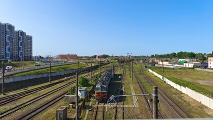 An electric locomotive stands on graveled and electrified railroad tracks. Behind a concrete fence are residential houses and a city street where cars drive. Sunny weather and blue sky
