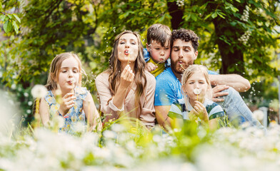 Fototapeta premium happy family sitting close together in high grass in spring blowing dandelion flowers