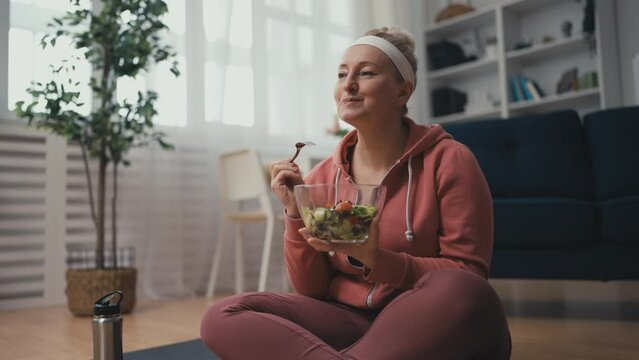 Middle aged woman in sportswear eating healthy salad after workout at home