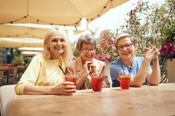 Mature ladies sitting in cafe terrace and talking while drinking cocktails and looking at camera