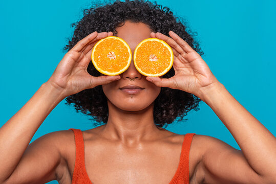Serious ethnic woman covering eyes with orange slices in blue studio