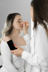 Young woman sits in comfortable chair beauty salon like professional makeup artist applies makeup to her face applying neat movements with each brush. Cosmetics and professional makeup artist services