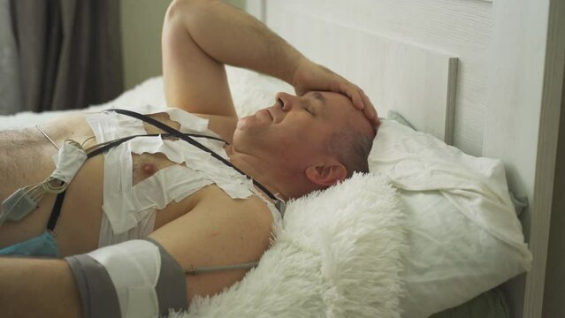 A man with a holter installed reveals malfunctions in the cardiovascular system. Lying on the bed, holding his head, rubbing his eyes.