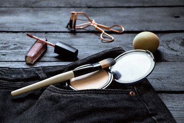 Cosmetics, powder and a brush lie in a jeans pocket, and lip gloss and a sponge lie side by side on...