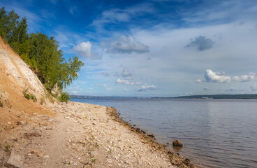 The bank of a large river with trees on the background of a blue sky with clouds in summer
