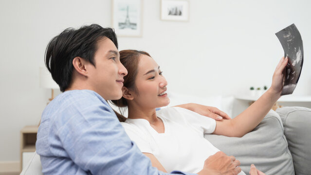 Asian pregnant wife holding ultrasound gift baby photo with young husband happiness and care lover while sitting on comfortable couch in living room at house