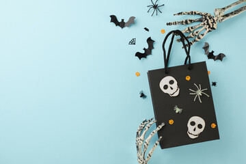 Halloween shopping atmosphere. Top view photo of creative package, skeleton hands, spooky decor on light blue background with promo spot