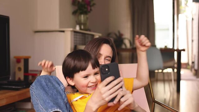 Happy mum have fun with son using smartphone apps at home, smiling mother hug preschool child boy having fun looking at cellphone relax with technology gadget, make video call, selfie. Mom kiss son.