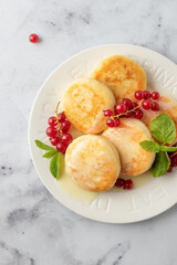 Cottage cheese pancakes or syrniki close up with red currant and milk sauce on white plate marble background top view, flat lay. Healthy tasty breakfast