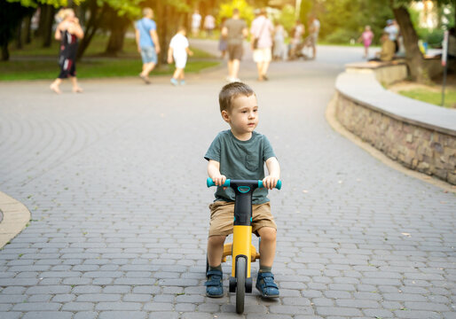 A Cute Toddler Boy Of Two Or Three Years Old Rides A Bicycle Or Balance Bike In The Park Against The Backdrop Of A Crowd Of People In The Background. Summer Activity For Children. Selective Focus