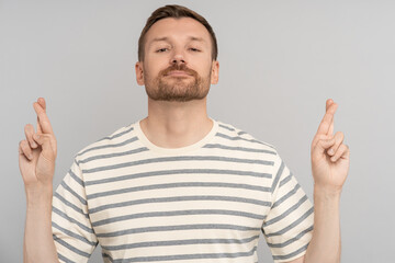 Naklejka premium Pleased man with fingers crossed waiting for onset of important moment, hopes for positive result, believes in luck, isolated studio background. Male shows with hands sign for good luck with desire