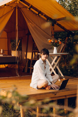 Beautiful woman in cozy outfit works at laptop while sitting on chair near the tent glamping