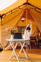 Beautiful woman in cozy outfit works at laptop while sitting on chair near the tent glamping