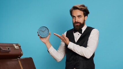 Doorkeeper shows wall clock on camera, checking time and trying to be punctual in hotel travel sector. Man hotel concierge using watch to see hour and minutes in studio next to trolley bags.