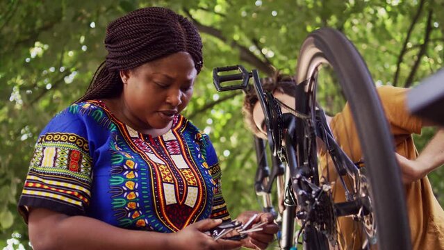 Young and sporty couple mending bicycle damages with specialized multitool in home yard. Healthy caucasian man and black woman selecting suitable allen key head for bike adjustments.