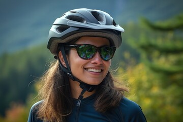 Fototapeta na wymiar A Middle Eastern mountain biker with her helmet and sunglasses sily fixed in place stands in a stand still close up portrait against a distant backdrop of forest green mountains.